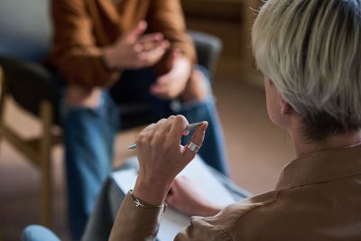 A therapist holding a pen speaks with a teenage client whose head is not visible.