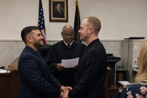 A same-sex couple wearing dark suits are married by a judge in a courtroom.