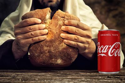 A figure representing Jesus breaks a loaf of bread with his hands on a rough wooden table while a can of Coca-Cola sits nearby