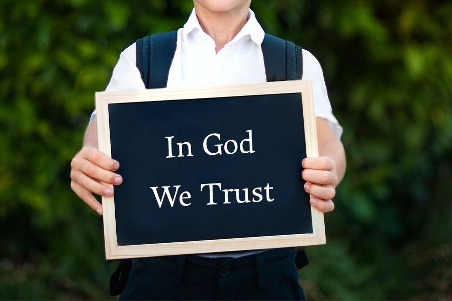 little boy in backpack holding sign that says in god we trust