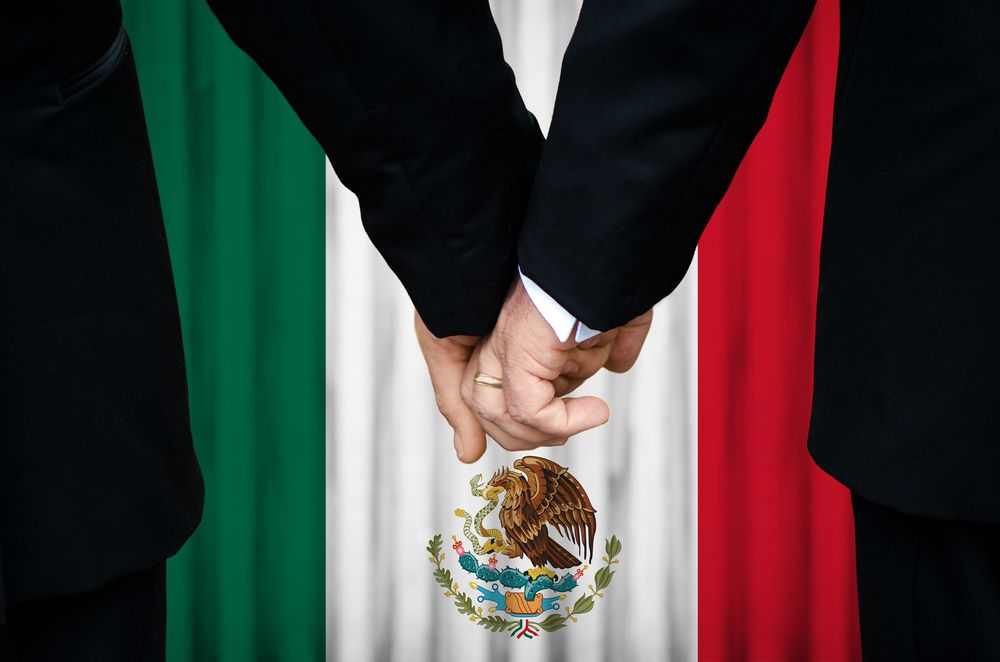 Two men holding hands in front of Mexican flag