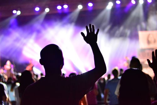 A silhouette of a man raising his hand in front of a crowd and a highly illuminated worship stage.