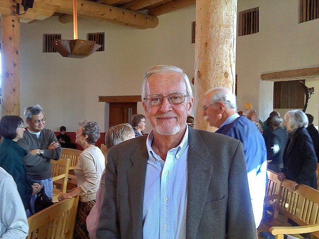 John Fife, wearing a grey blazer and a blue shirt, smiles at the camera from the inside of his crowded Tuscon church.