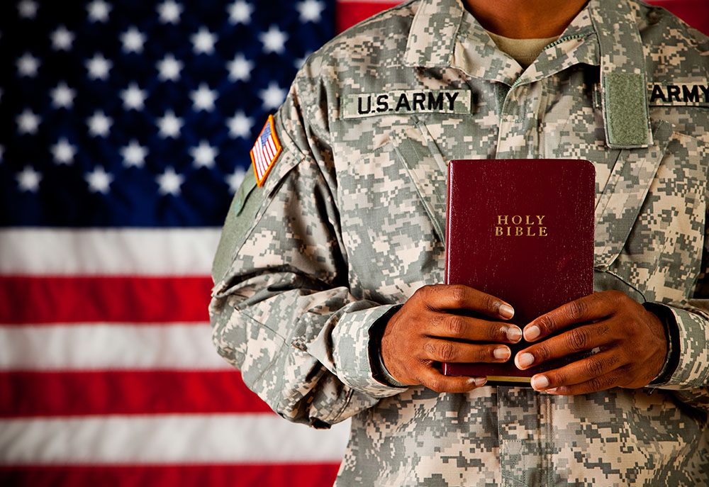 A soldier holds a Christian bible while standing in front of a US flag.
