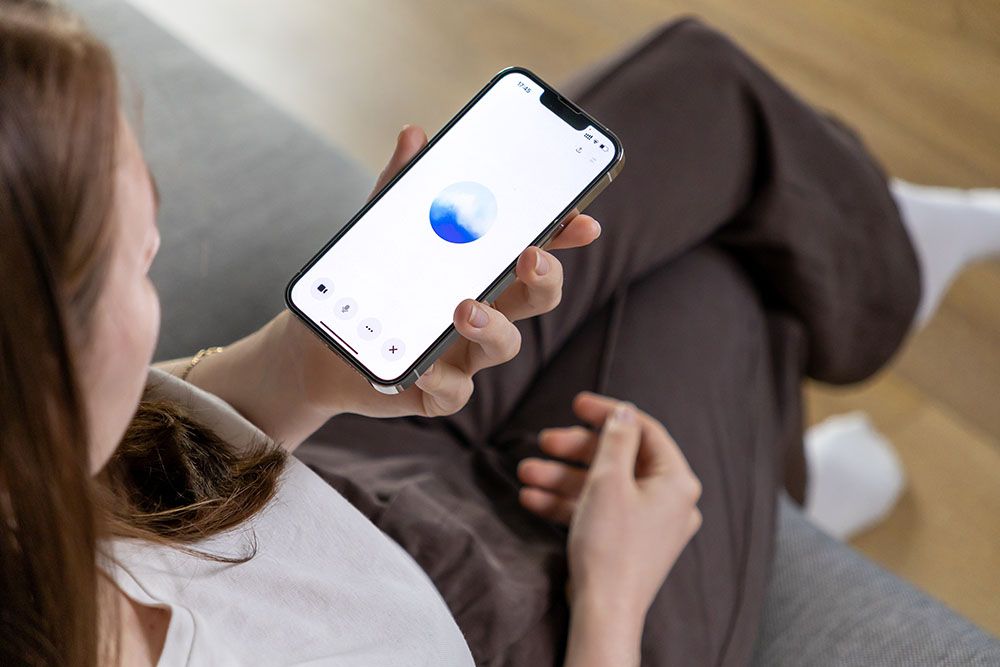 A woman in a white top and brown trousers sits on a sofa and talks to the ChatGPT chatbot on her mobile phone.