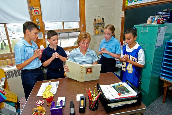A teacher reads from the Bible to a group of students at her desk.