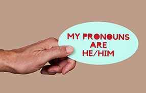 A hand holds a sign reading "my pronouns are he/him"