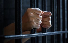 A man's hands clutch prison bars in a dark cell